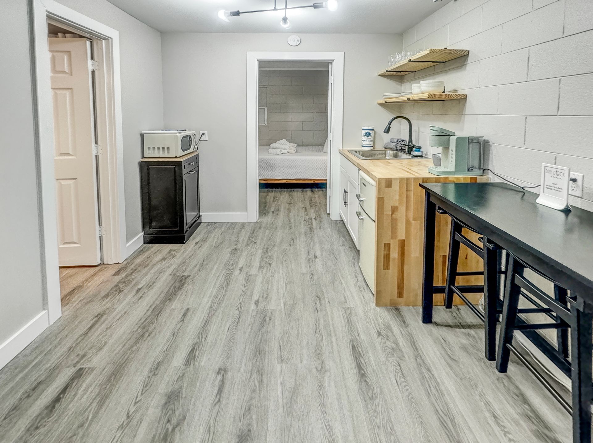 A small kitchen area with a microwave, sink, and table. Gray flooring and walls. A bedroom is visible in the doorway.