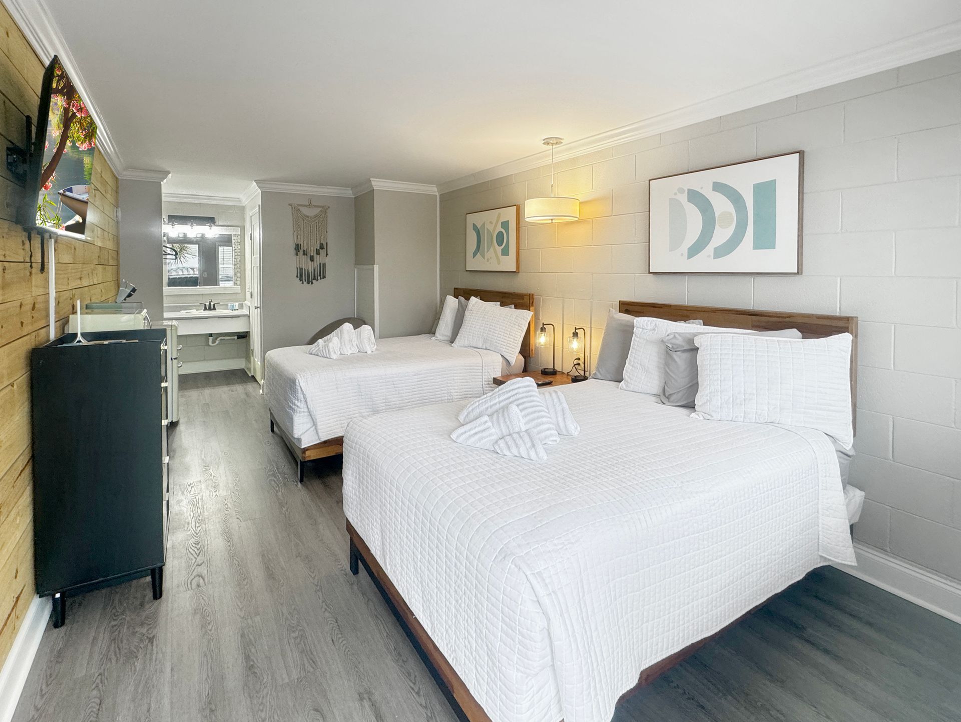 A hotel room with two queen beds covered in white bedding. Gray walls, light wood floors, and a dresser are visible.
