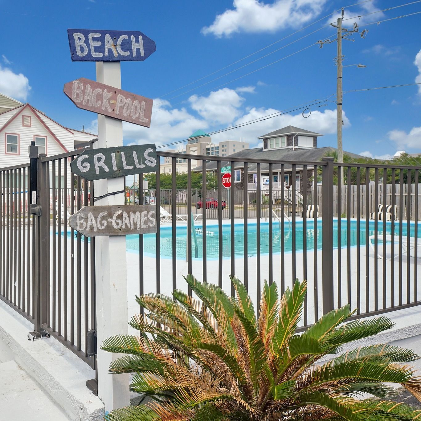 Signpost directs to beach, back pool, grills, and bag games near a fenced swimming pool. Blue sky, sunny day.