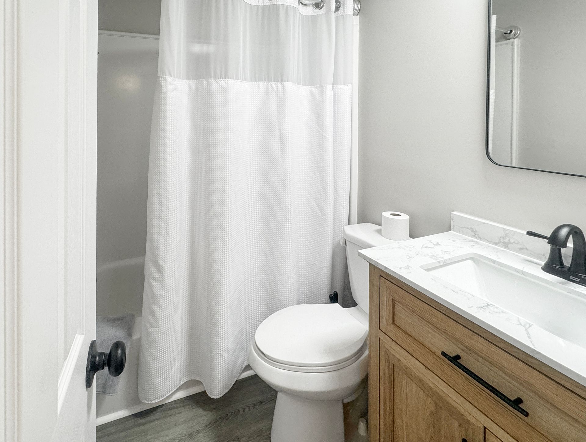 A small, bright bathroom with a tub and shower, toilet, vanity with a sink, and a closed door. White shower curtain.
