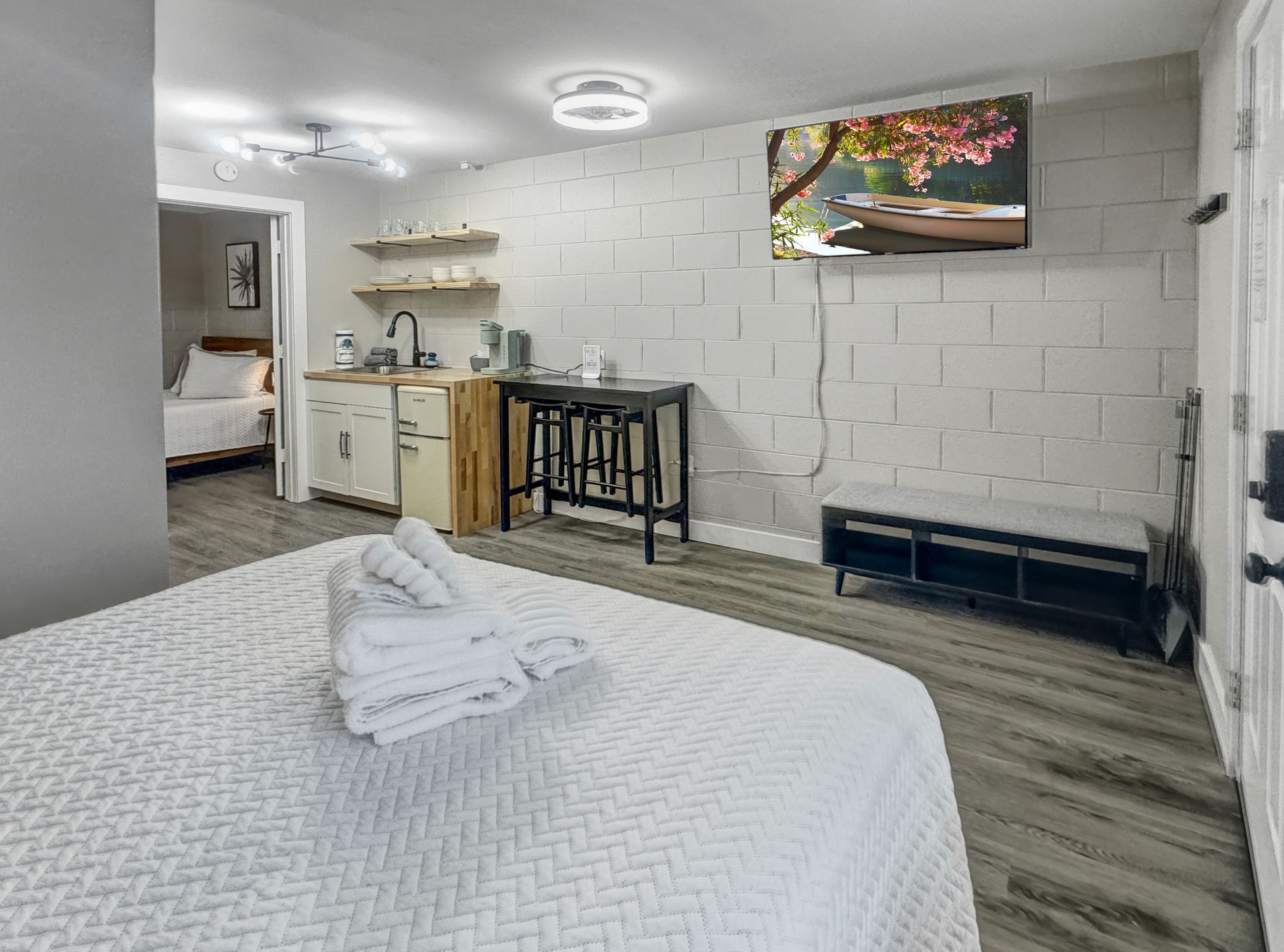 A hotel room with a bed, kitchenette, and TV. Light gray walls, a small table with stools, and a view into the bedroom.