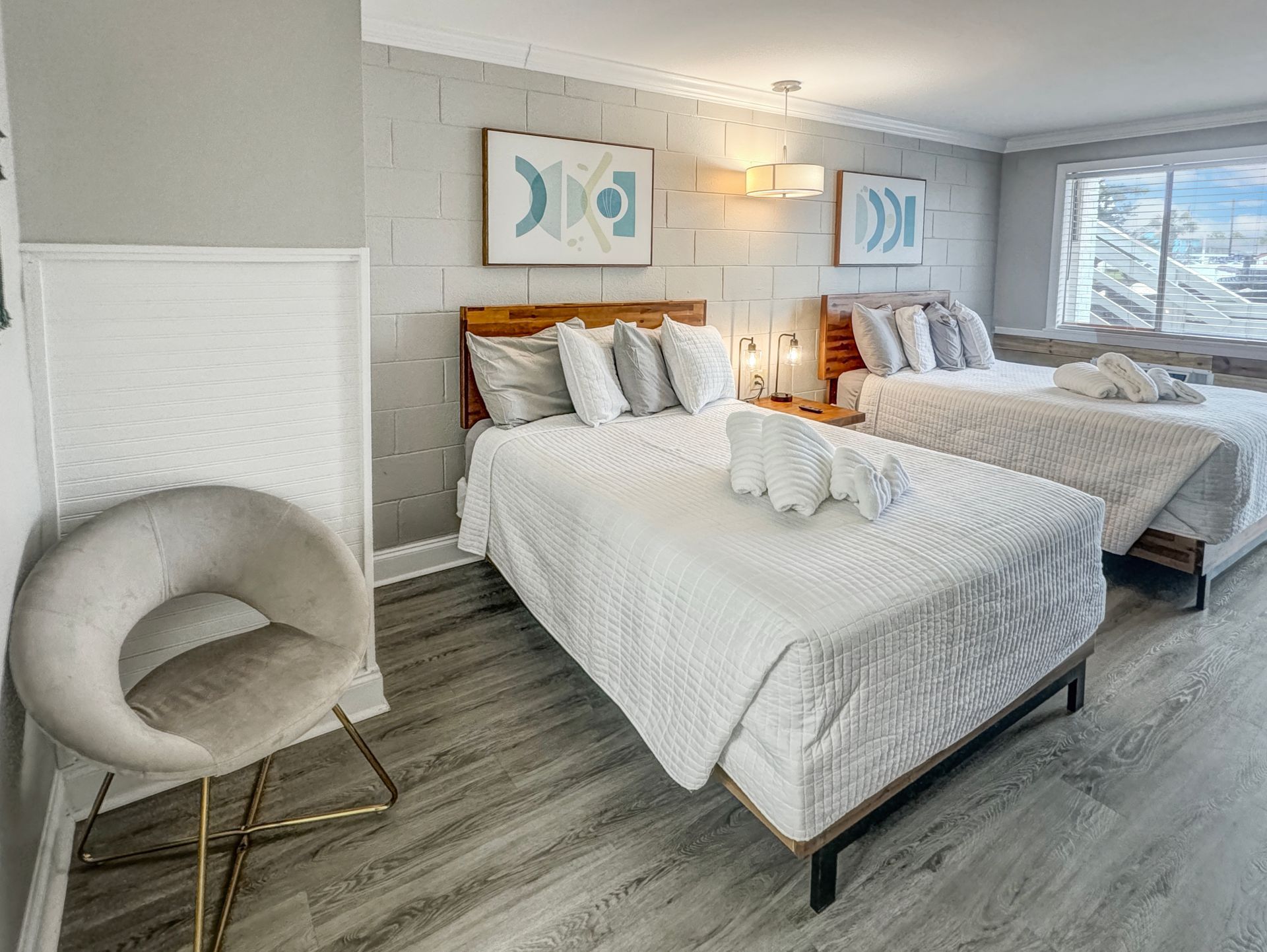 A modern bedroom with two beds, light gray walls, a gold chair, and artwork. Wooden headboards and white bedding.
