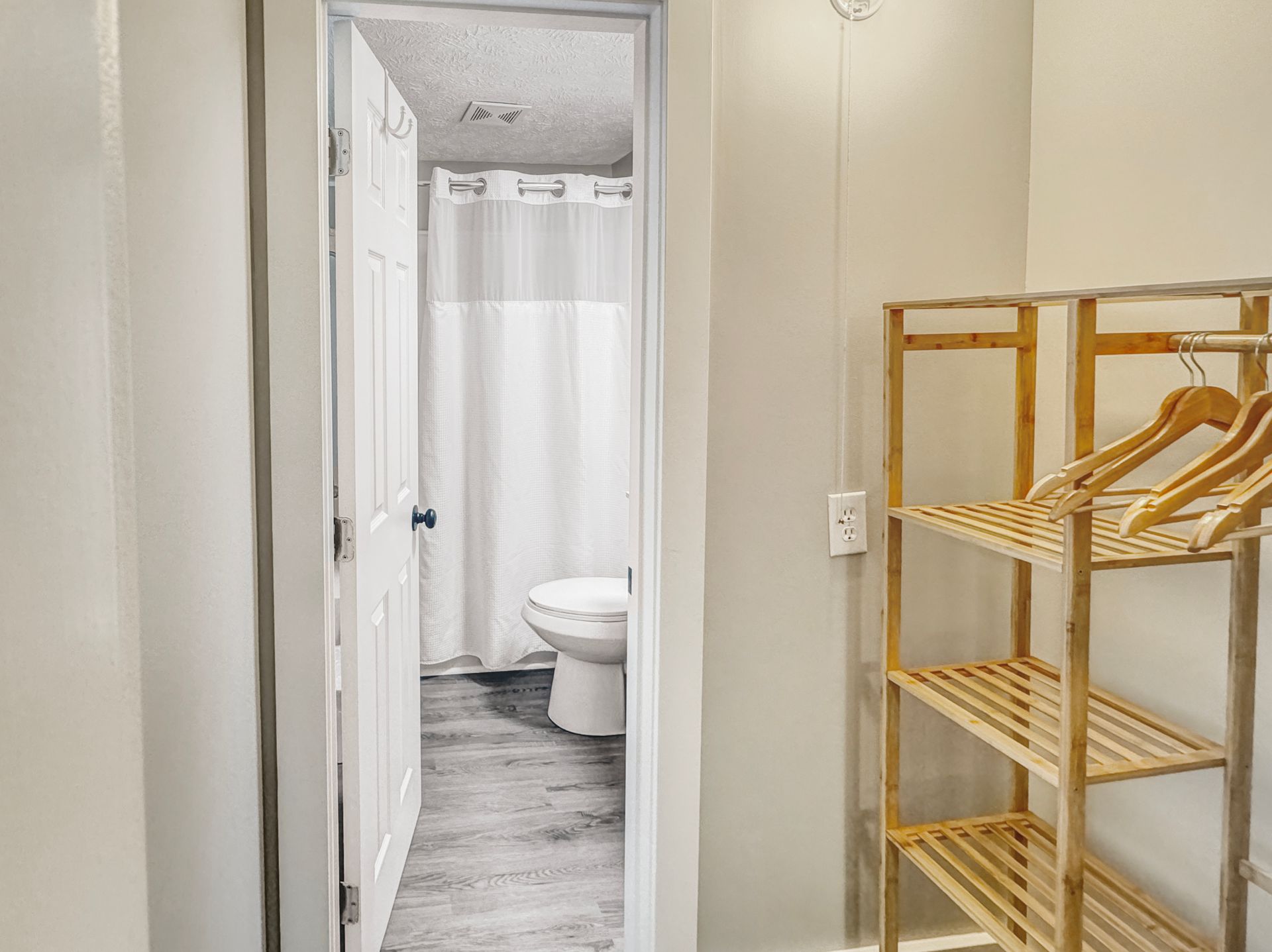 Open doorway into a bathroom with a toilet and shower. A wooden rack with hangers stands on the right.