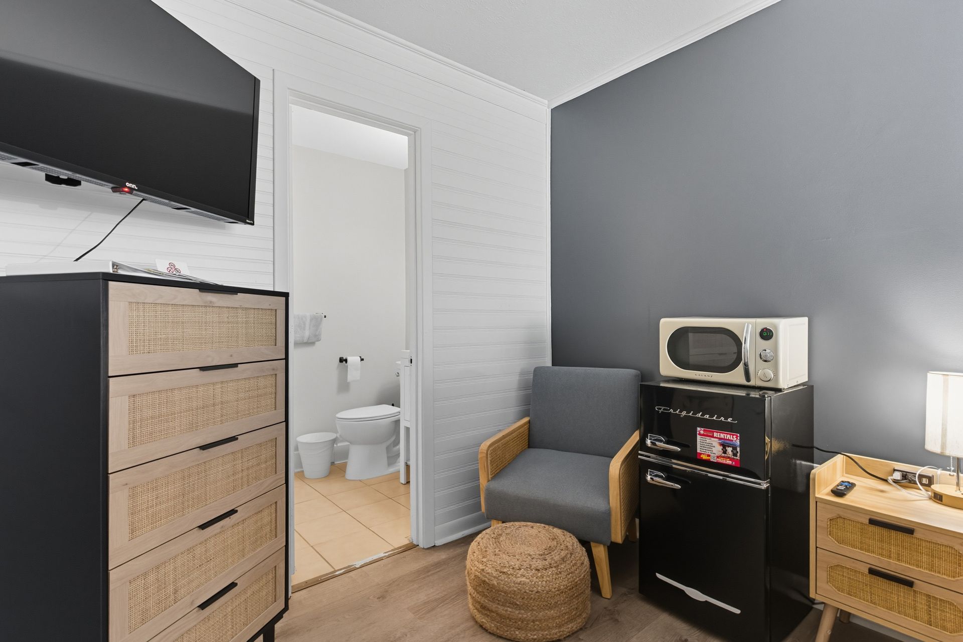 Small room with a TV, dresser, chair, mini-fridge, microwave, nightstand, and doorway to a bathroom.