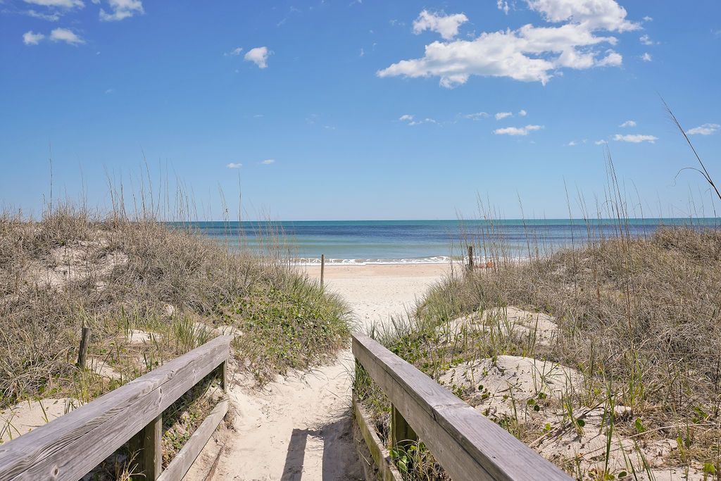Wooden walkway leading to a sandy beach with blue ocean and sky, flanked by grassy dunes.