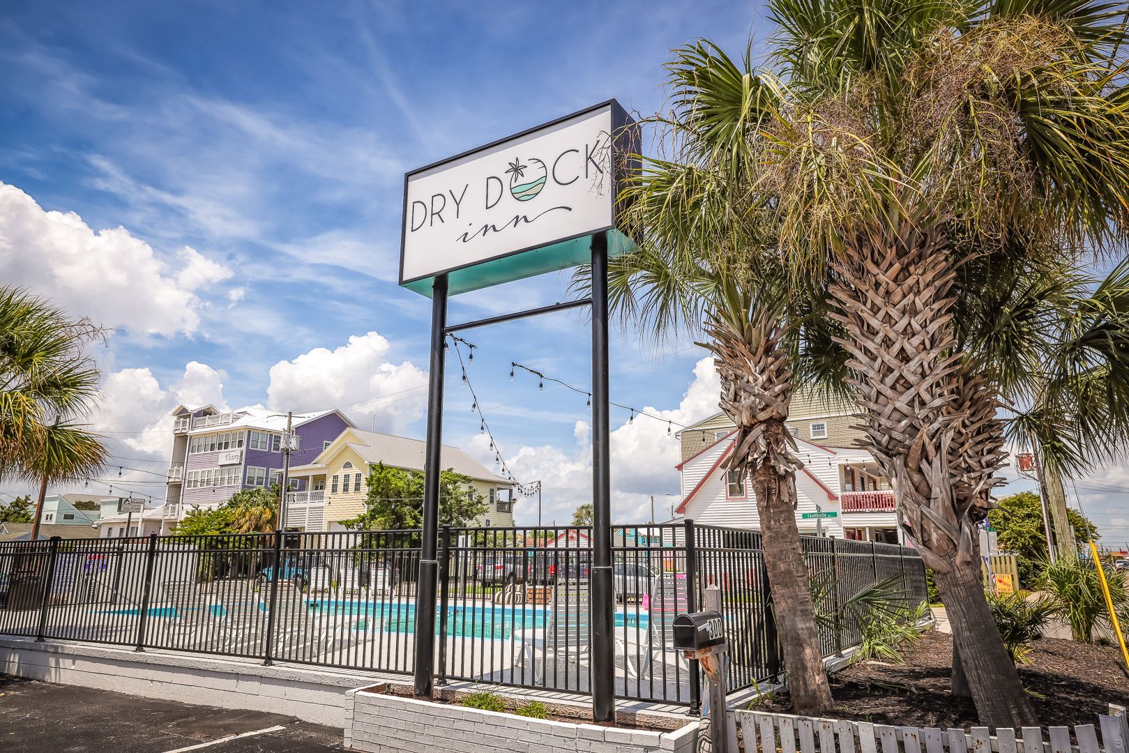 Sign for Dry Dock Inn with pool behind a fence, palm trees, and colorful buildings.