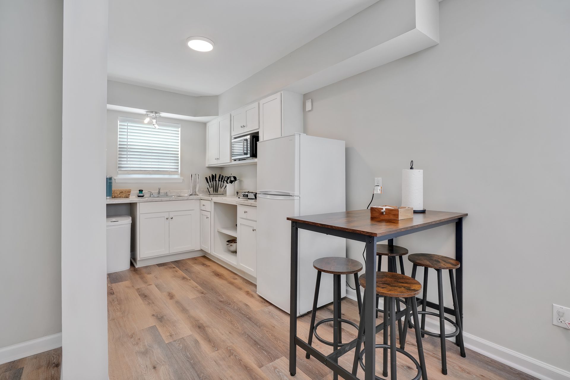 Small kitchen with white cabinets, appliances, and a small table with stools; wood flooring.