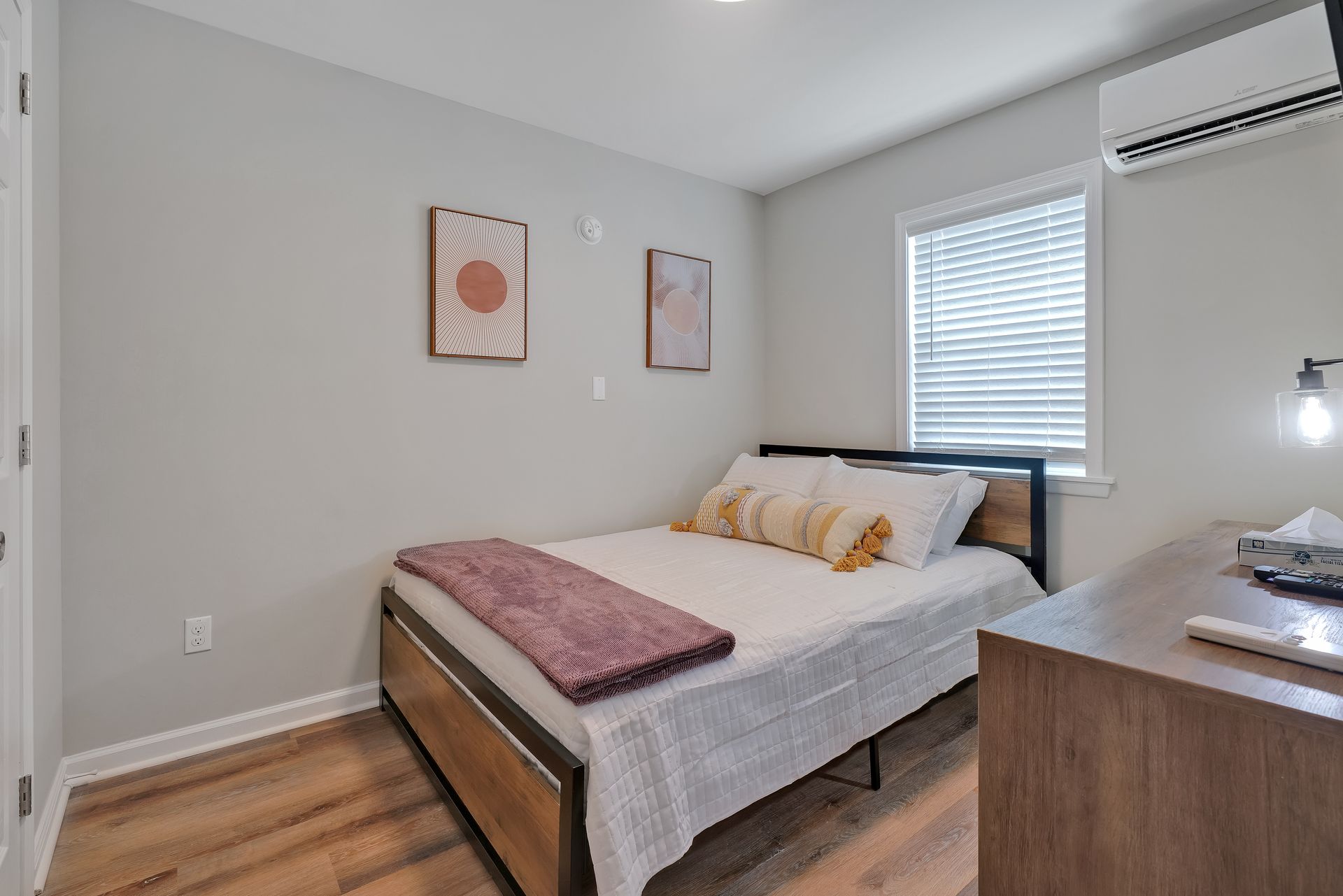 Bedroom with a bed, desk, and air conditioner; light gray walls, wood-look floor, and simple artwork.