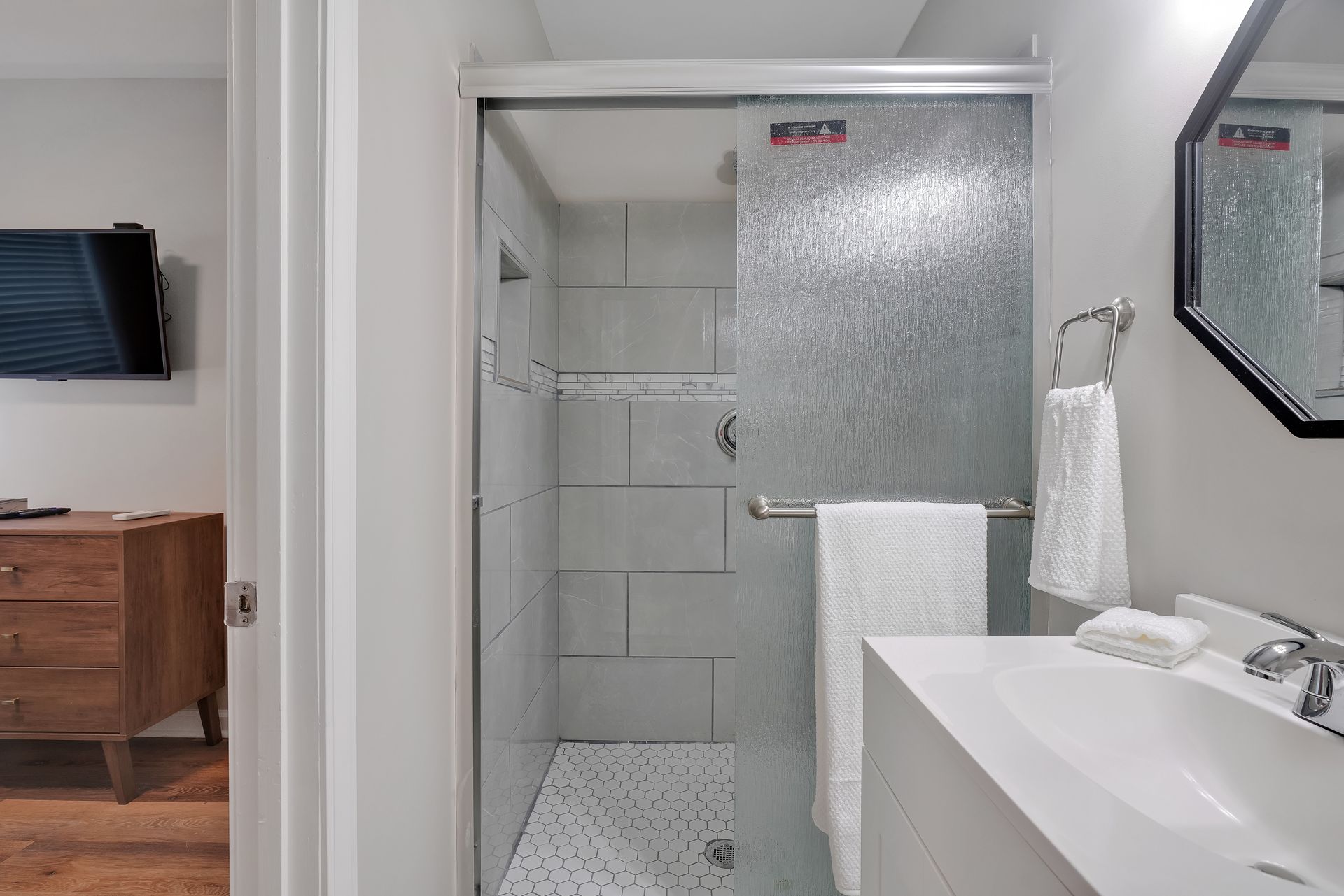 Bathroom with a glass shower, white sink, towel rack, and a dresser visible in the doorway.