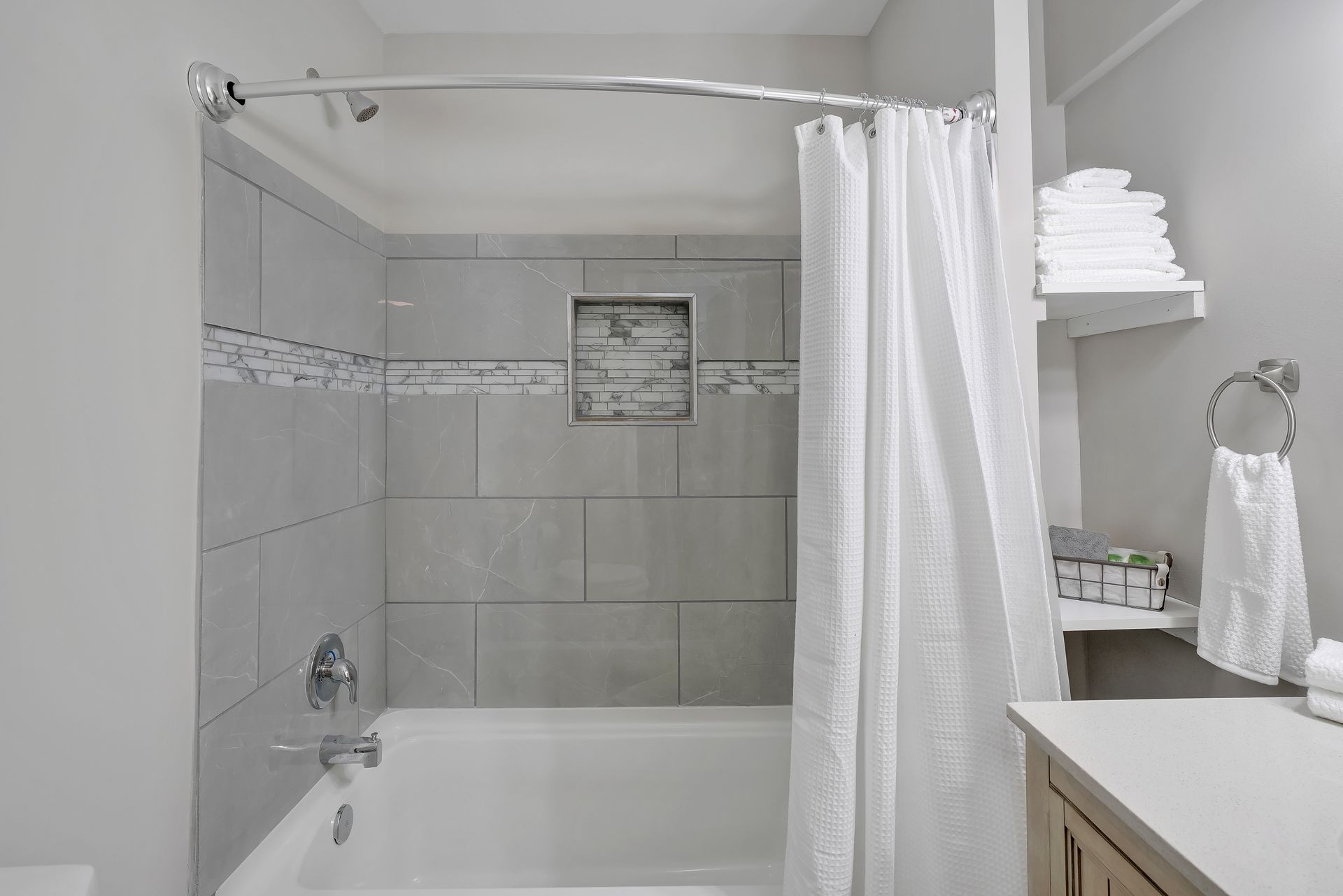 Bathroom with a white bathtub, gray tiled walls, shower curtain, and a shelf with towels.
