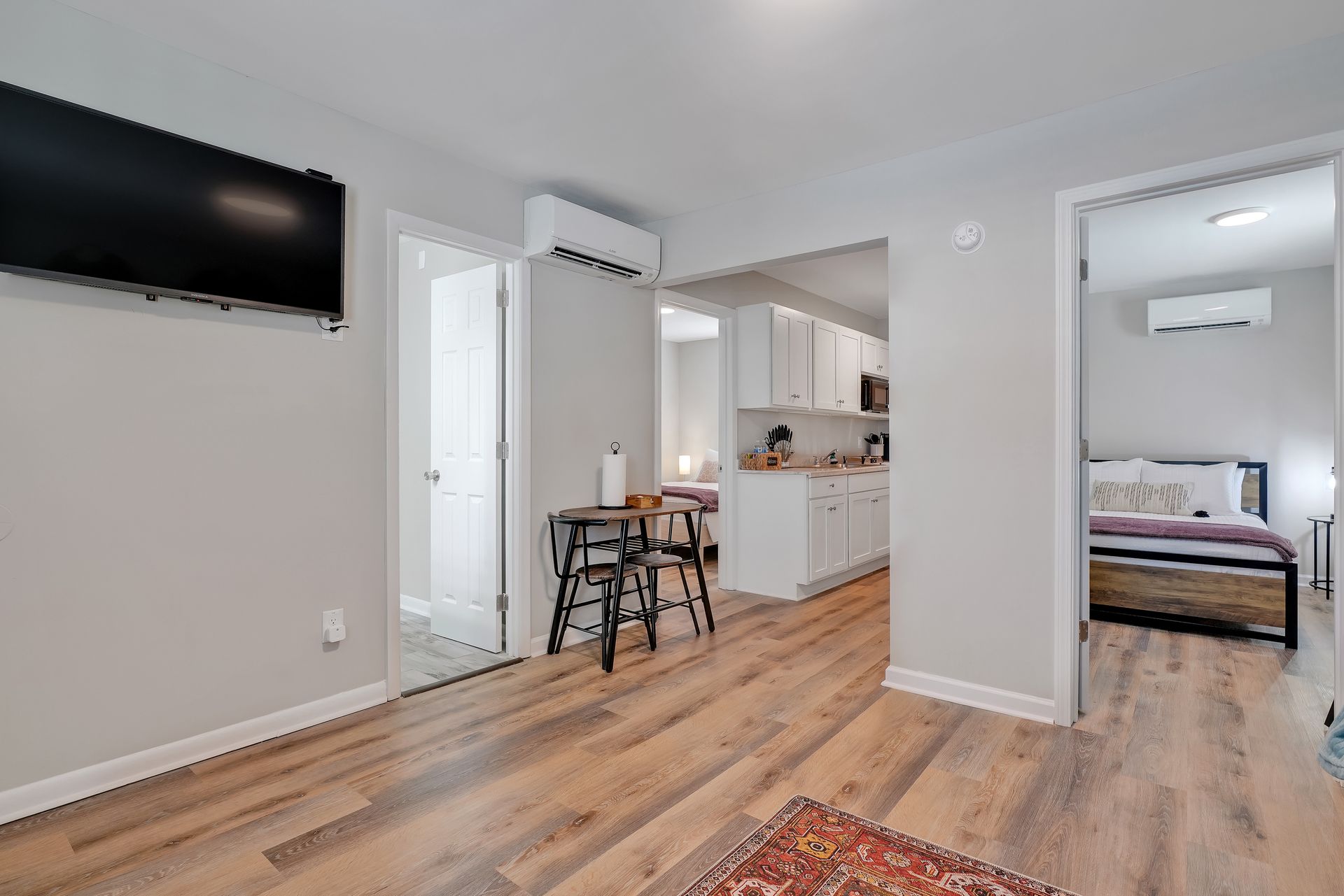 Small apartment with light wood floors, open to kitchen and bedroom, with wall-mounted TV and air conditioners.