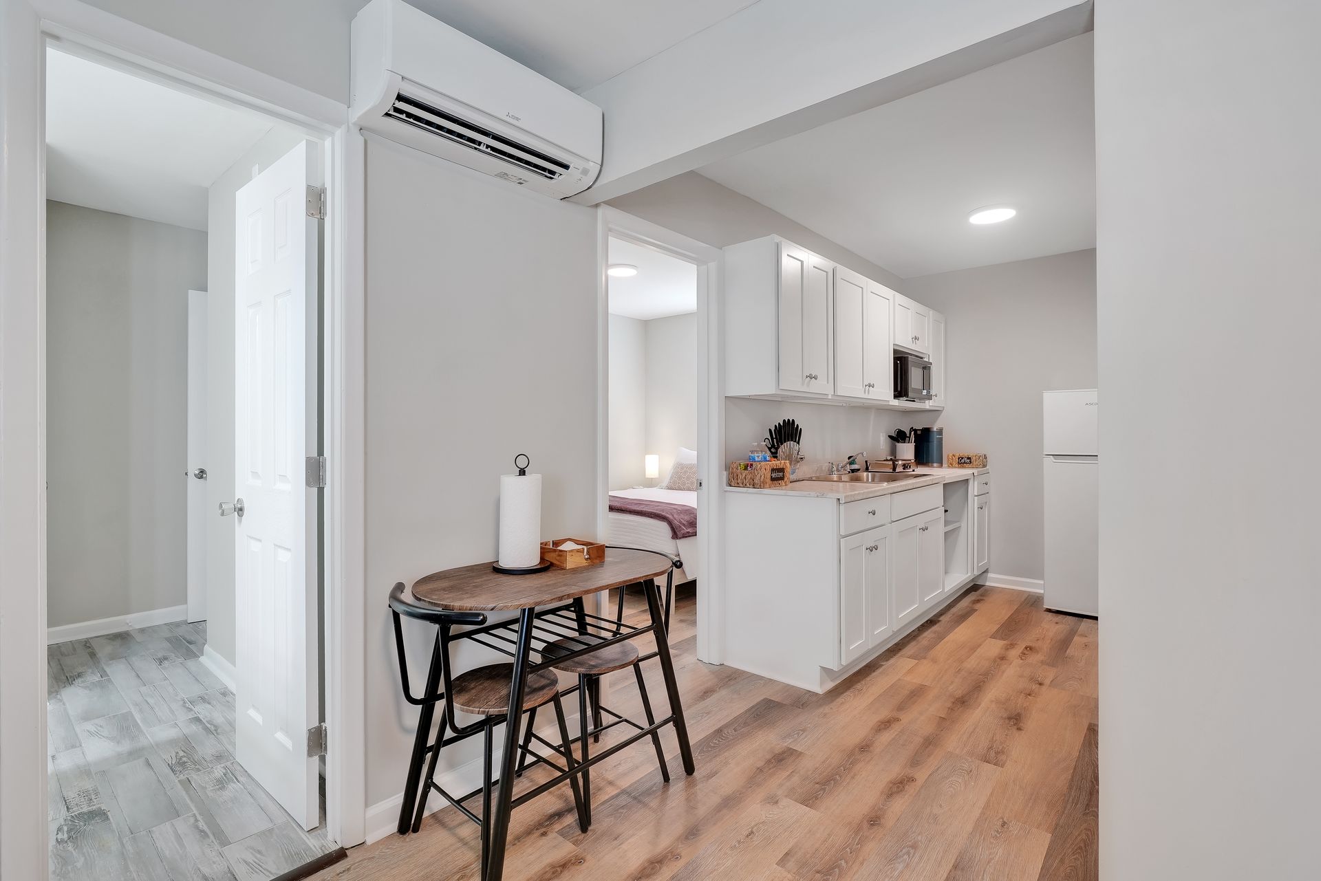 Interior of a small apartment with a kitchen, dining area, and bedroom. White cabinets, light wood floors.