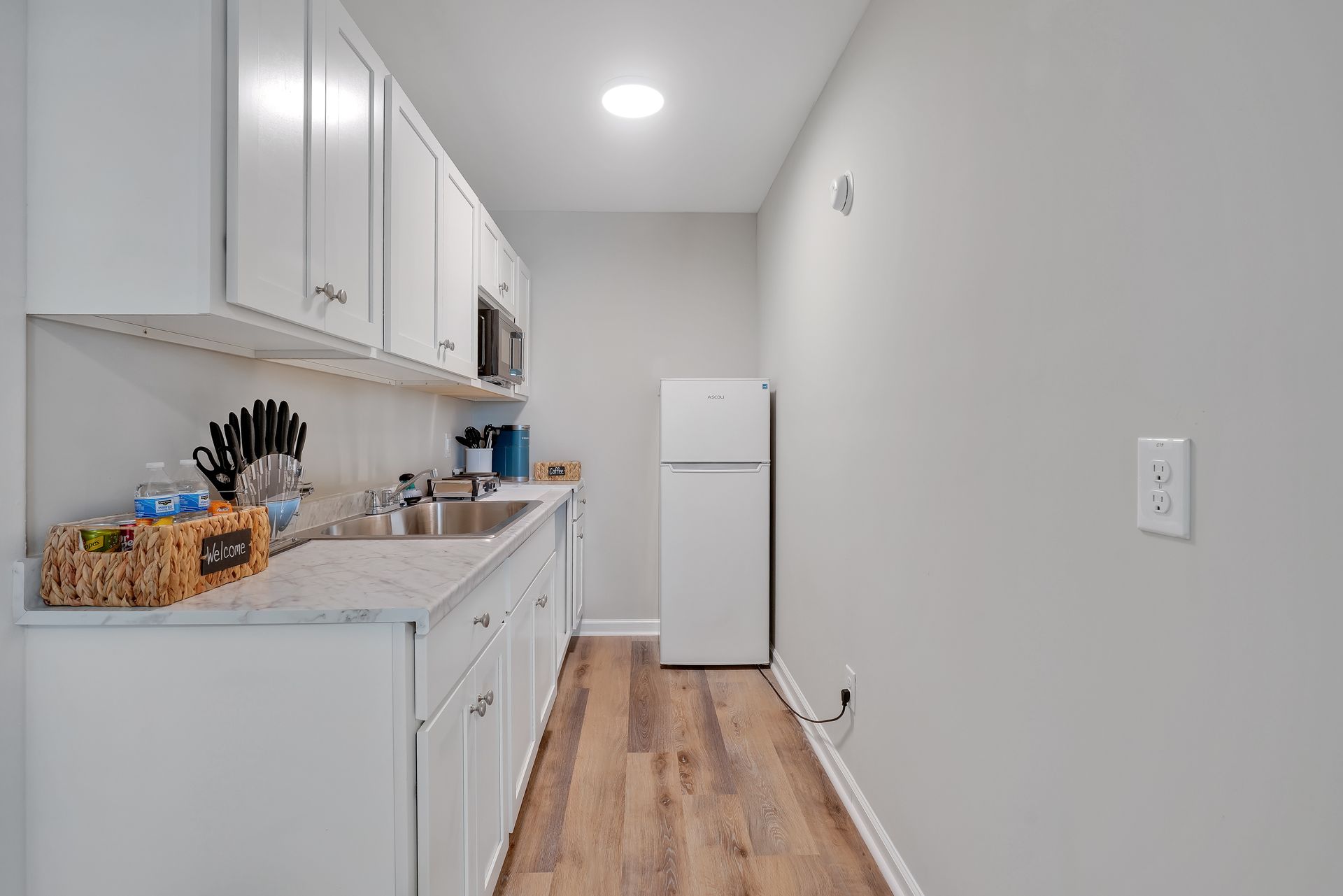 A small, white galley kitchen with cabinets, appliances, and a wood-look floor.