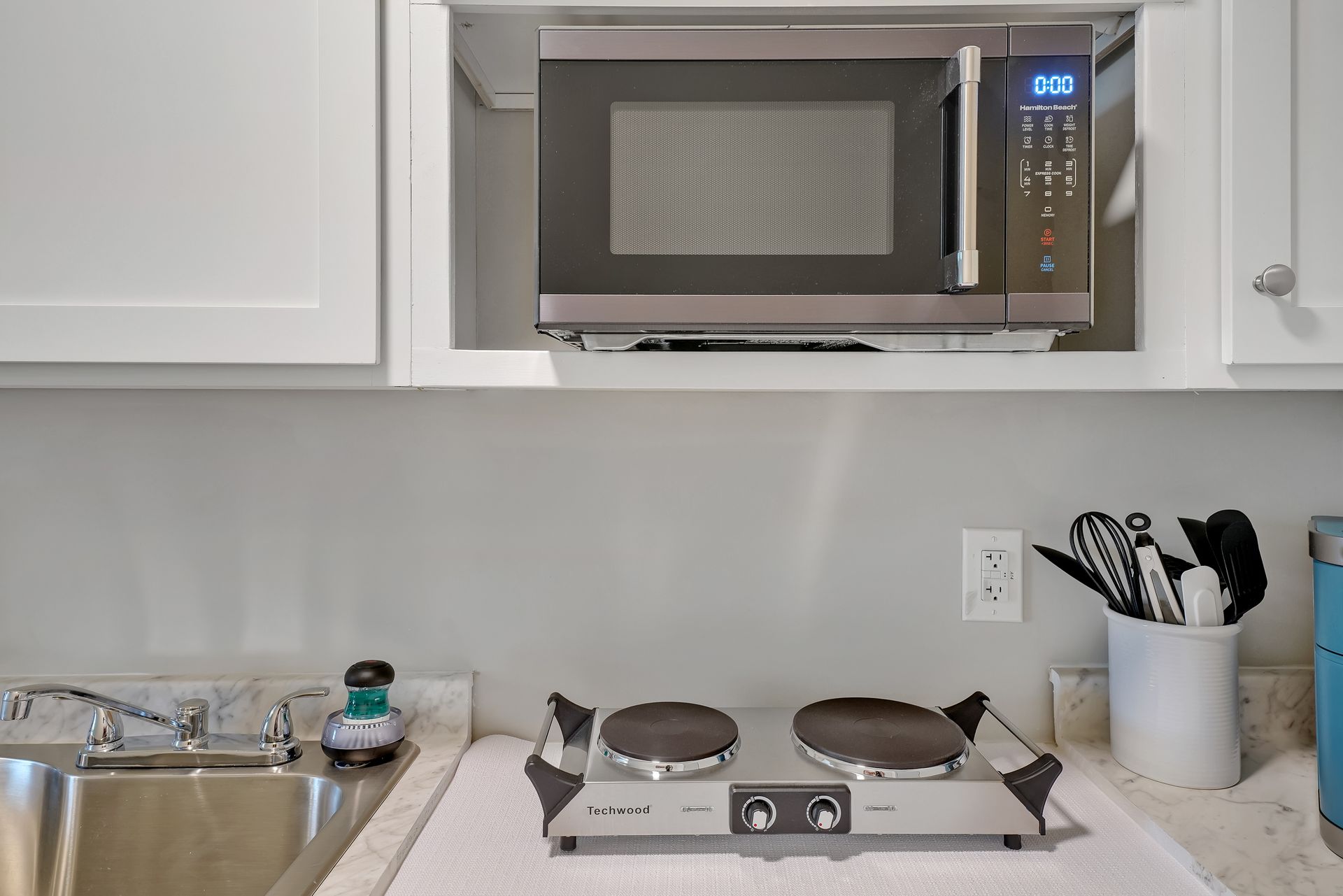 Microwave above a portable two-burner cooktop on a countertop with a sink and utensils.