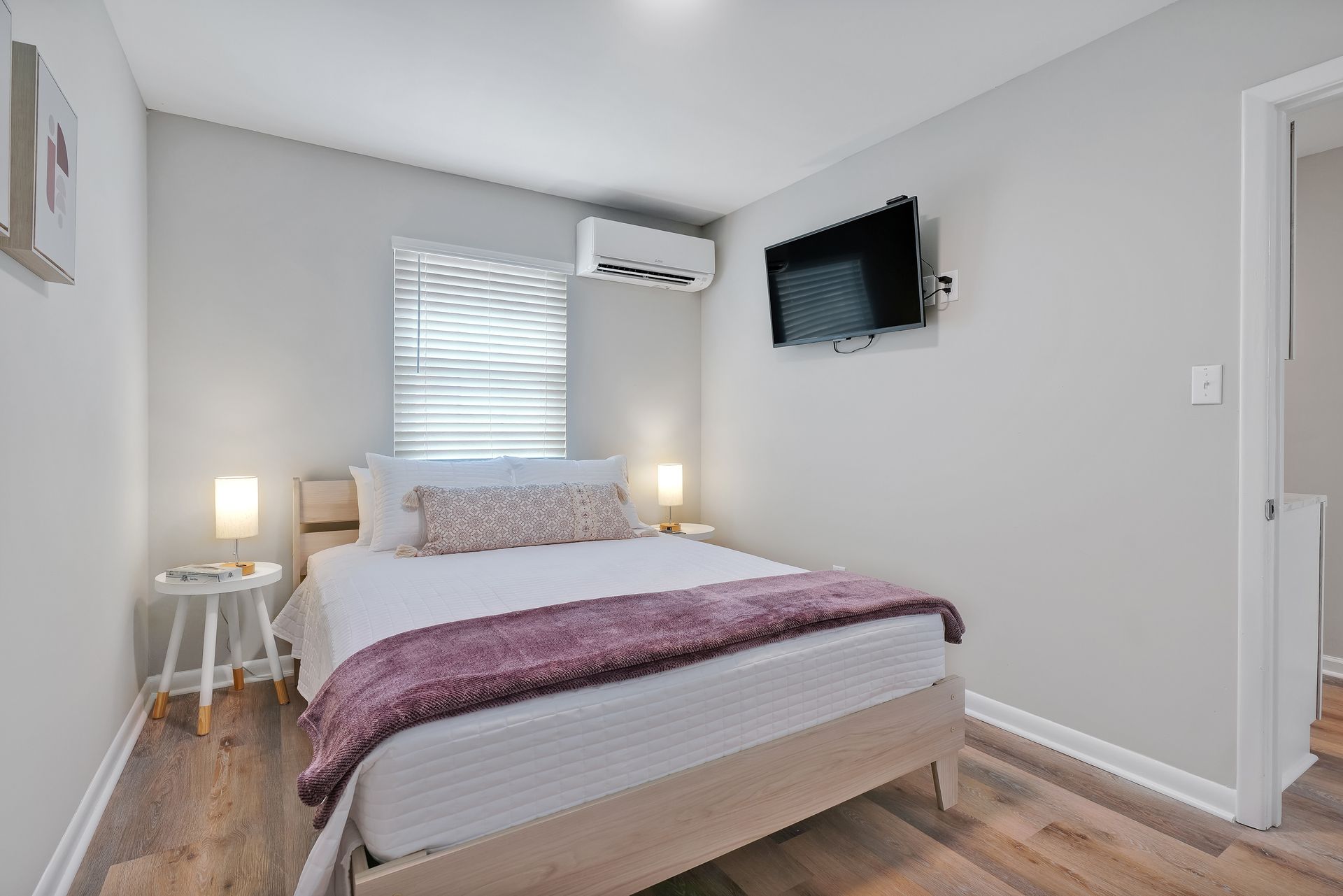 Bedroom with bed, TV on wall, and small table. Light gray walls, light wood floor.
