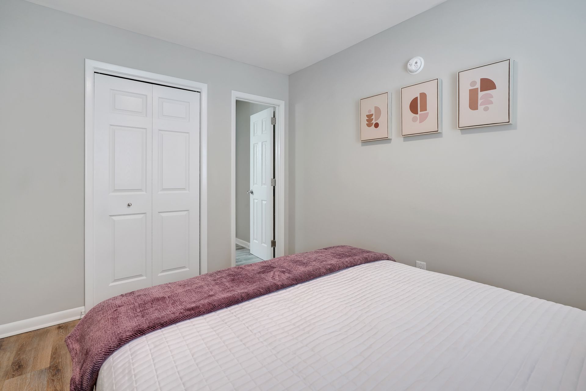 Bedroom with white bedding, purple throw, artwork on gray wall, and white closet door.