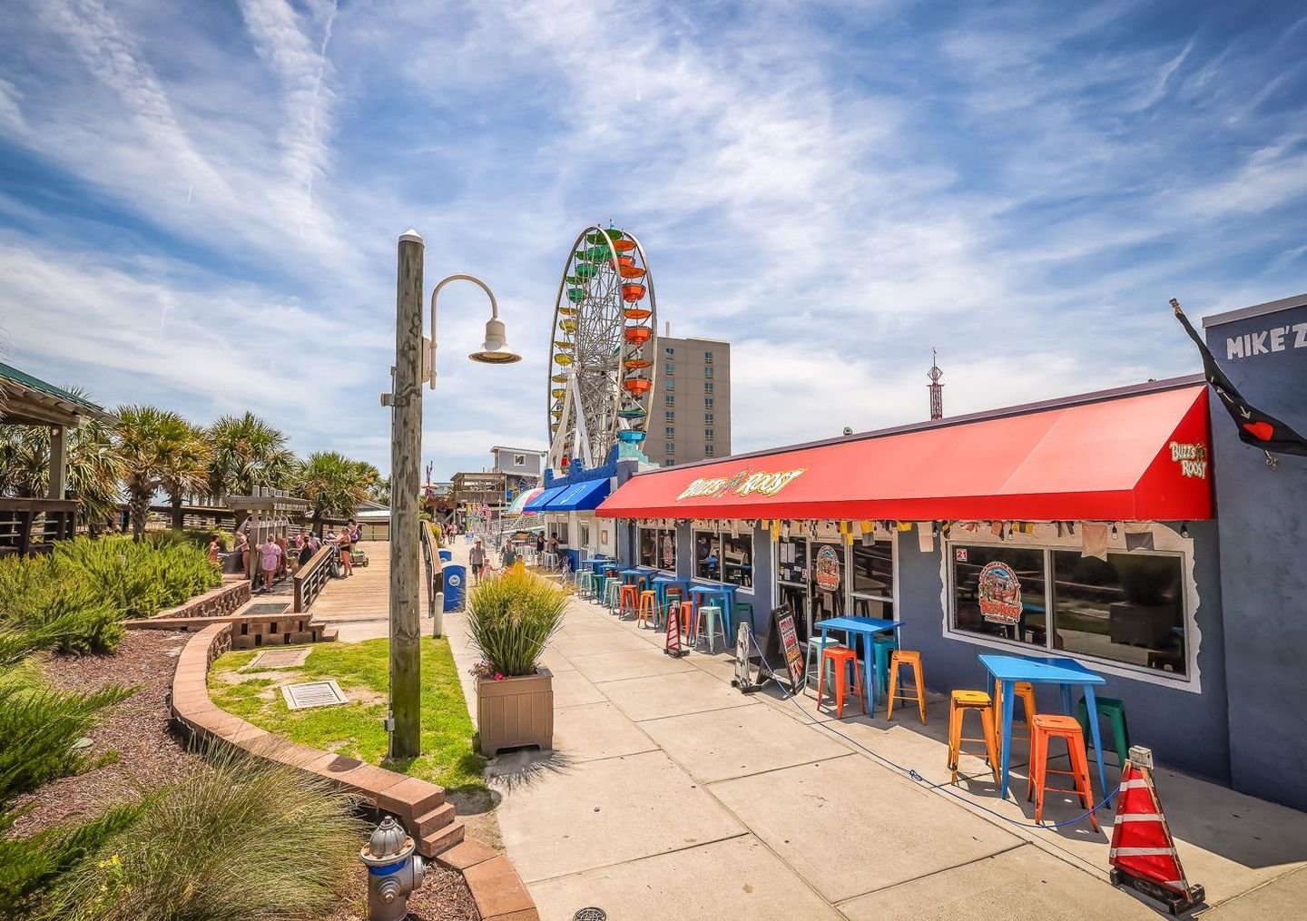 A seaside boardwalk with a colorful Ferris wheel, shops, and outdoor seating under a blue sky.