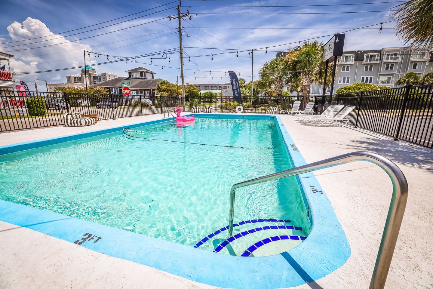 Swimming pool with a flamingo floatie; surrounded by a fence, patio, and buildings. Bright sunny day with a clear sky.
