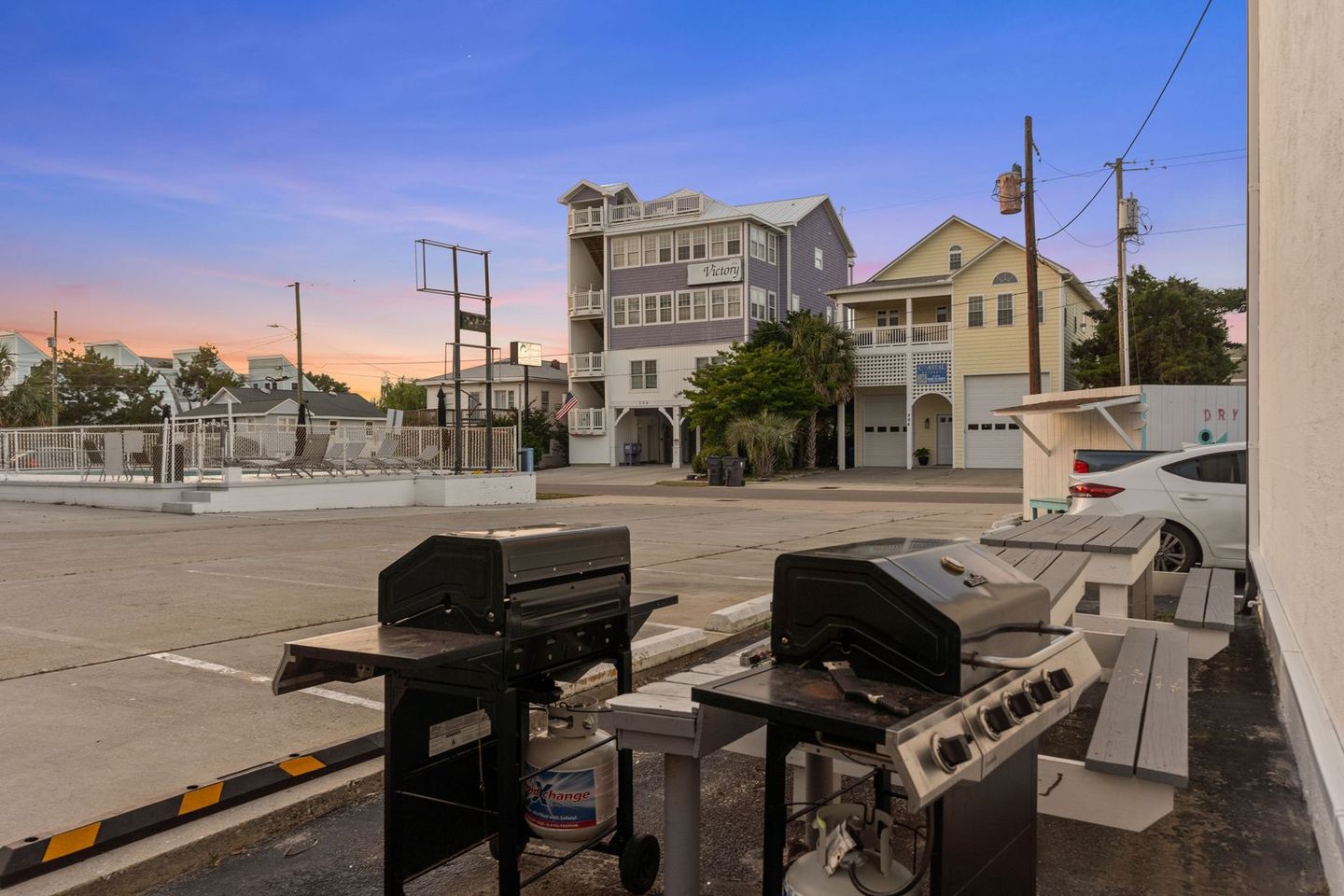 Two outdoor grills on a patio, with buildings and a parking lot in the background under a colorful sunset sky.