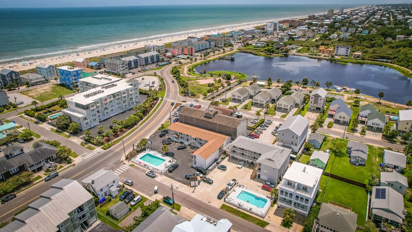 Aerial view of a coastal town with a beach, ocean, lake, and buildings, featuring a hotel with a pool.