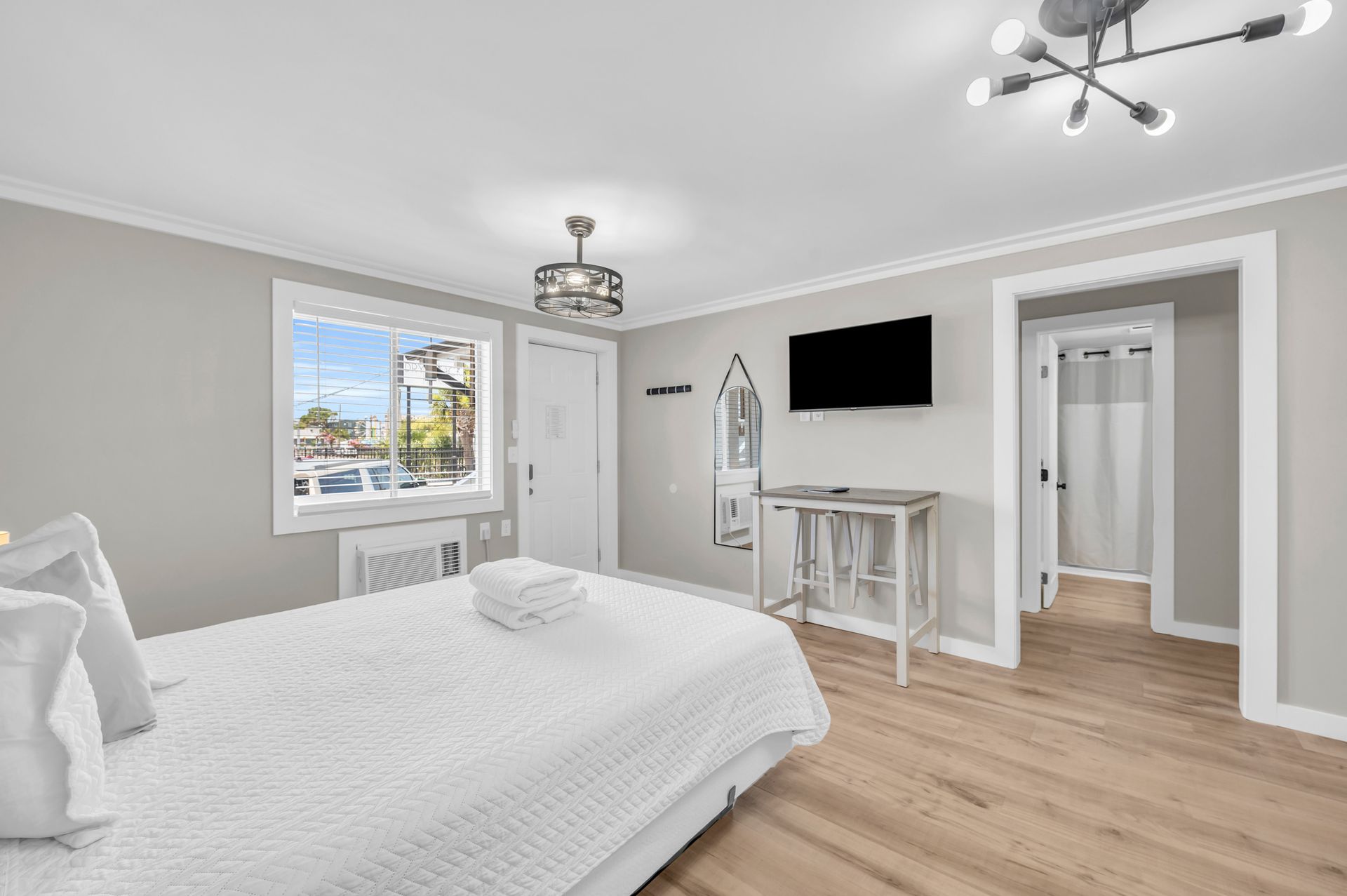 A bedroom with a bed, TV, and doorway. The room is light gray with light wood floors and white trim.
