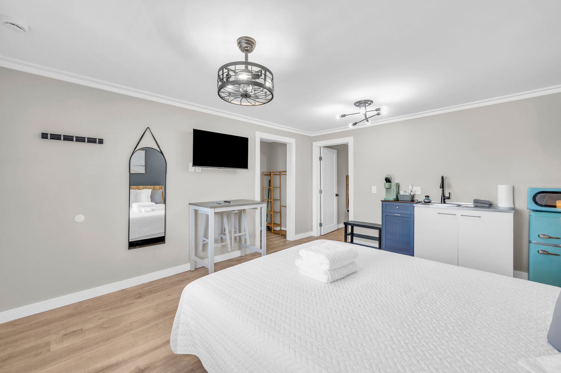 Bedroom with a large bed, a mirror, a TV, and a small kitchen area. Gray walls, wooden floors, and neutral colors.