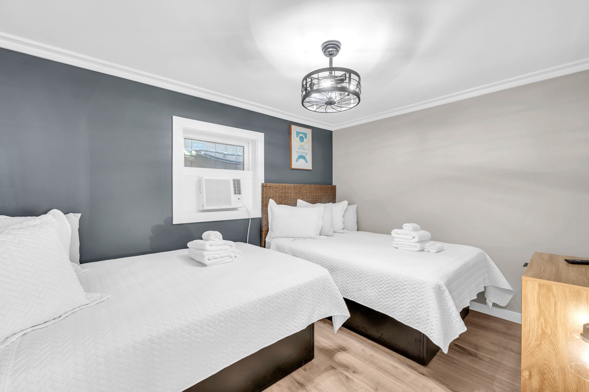 Two twin beds in a bedroom with white bedding, gray and light gray walls, and a small window with an air conditioner.