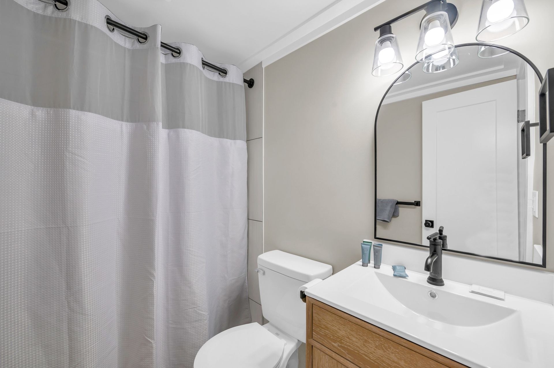 Bathroom with a gray and white shower curtain, white toilet, wooden vanity, and arched mirror.