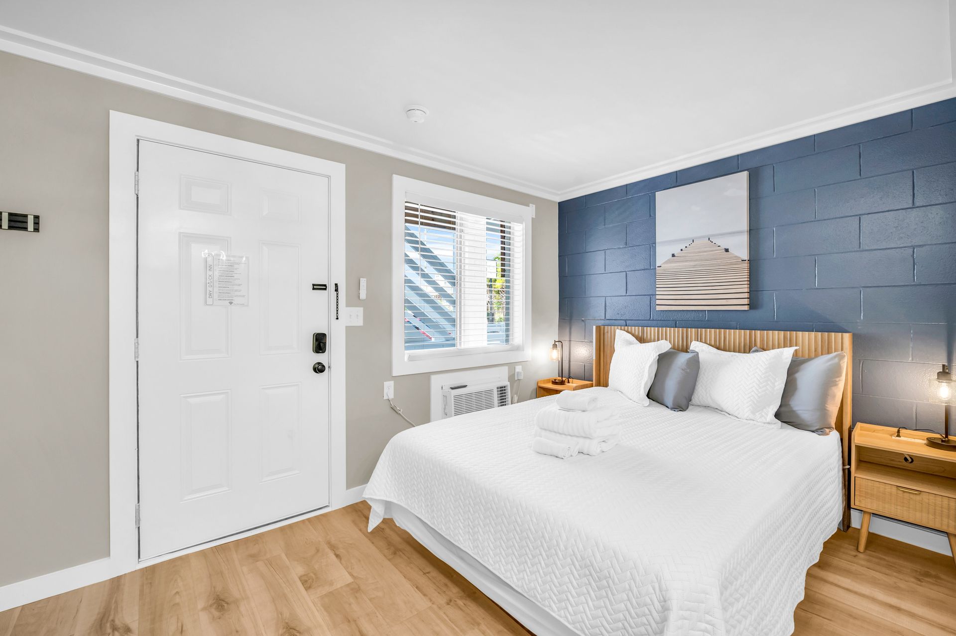 Bedroom with a white bed, blue accent wall, and wooden nightstands. The room has a door, window, and artwork.