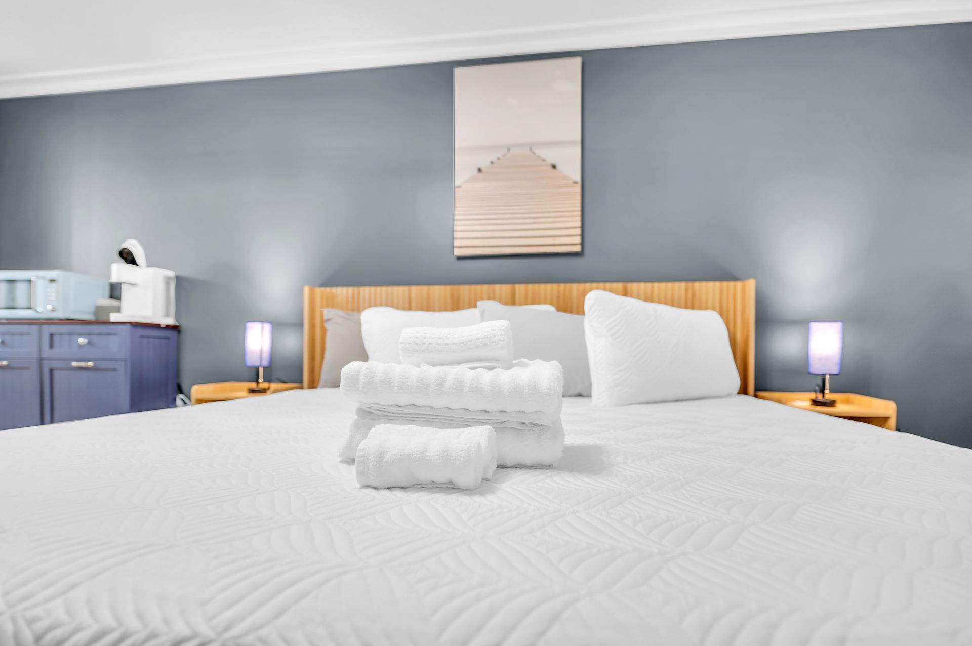 A bedroom with a large bed, towels, and a painting. The walls are grey, and there are nightstands on either side of the bed.