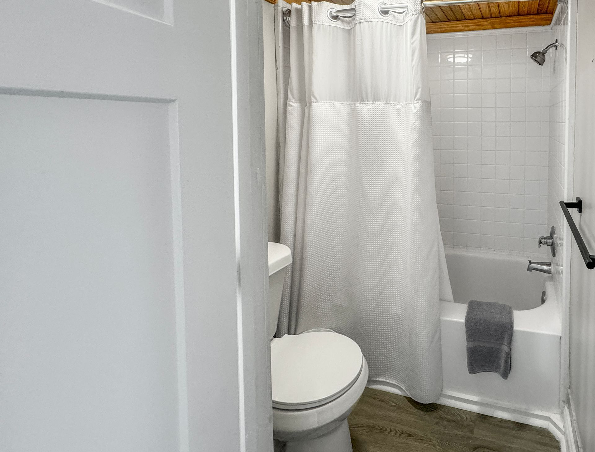 A small bathroom with a white tub and toilet, white tiled walls, a gray towel, and a white shower curtain.