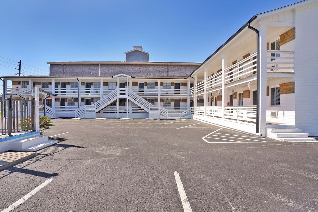 Exterior view of a white two-story motel with balconies, stairs, and a parking area. Clear blue sky overhead.