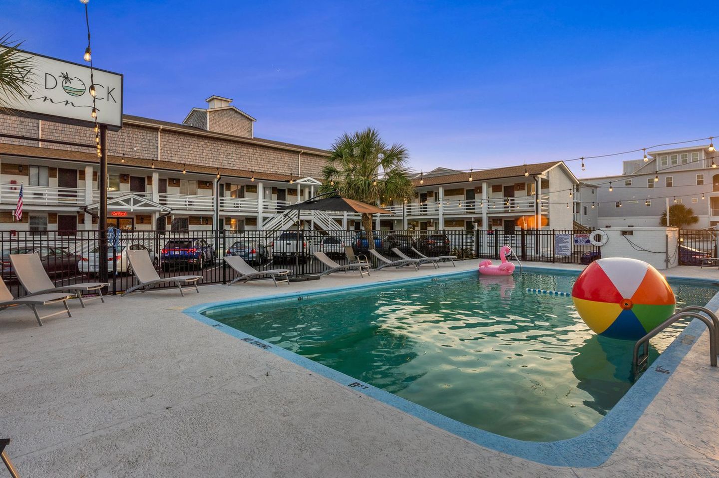A motel pool at dusk, with a large beach ball and a pink flamingo float. Lounge chairs and the motel buildings are visible.
