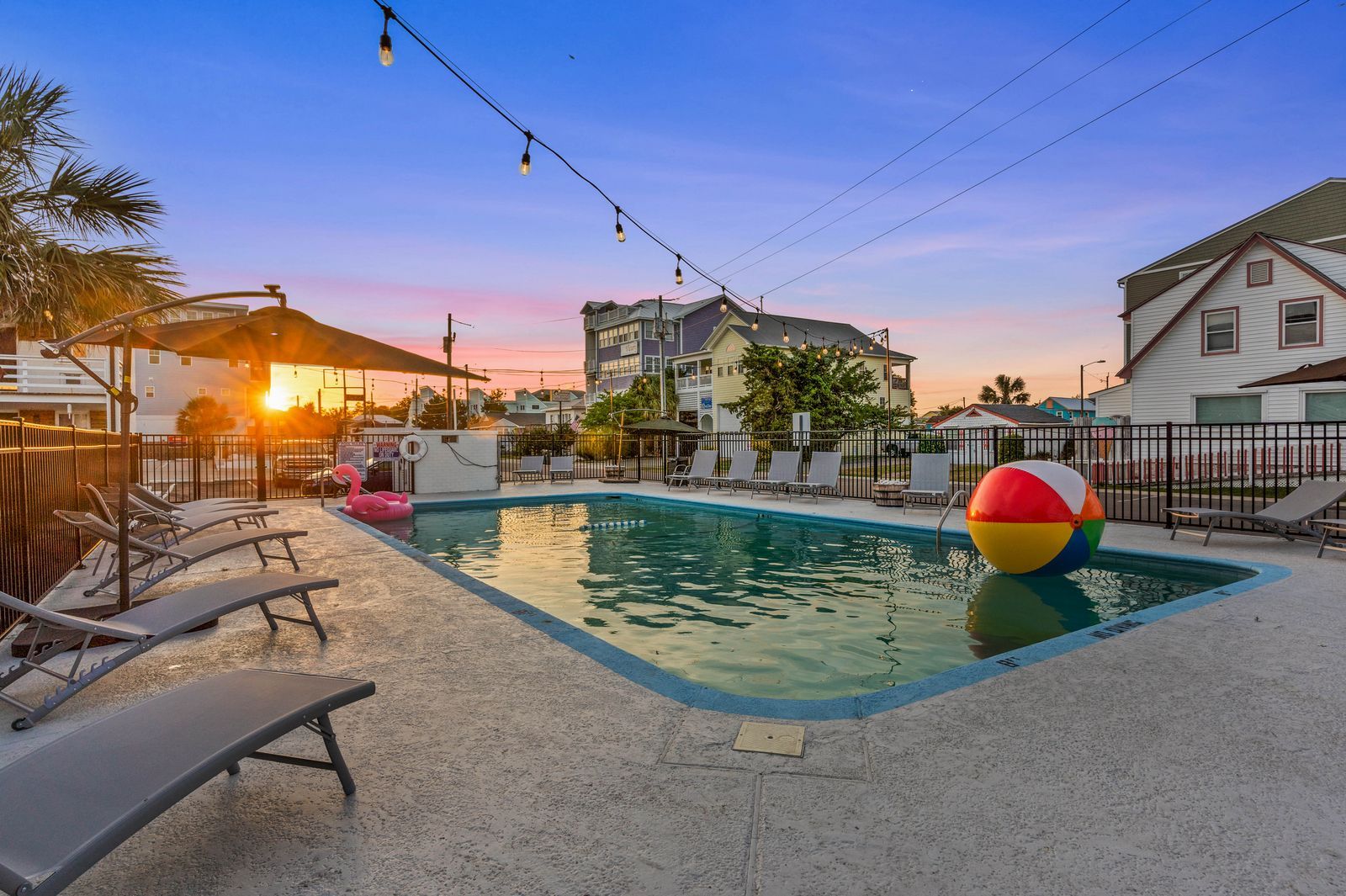A pool area at sunset with lounge chairs, string lights, and a large beach ball. Buildings are visible in the background.