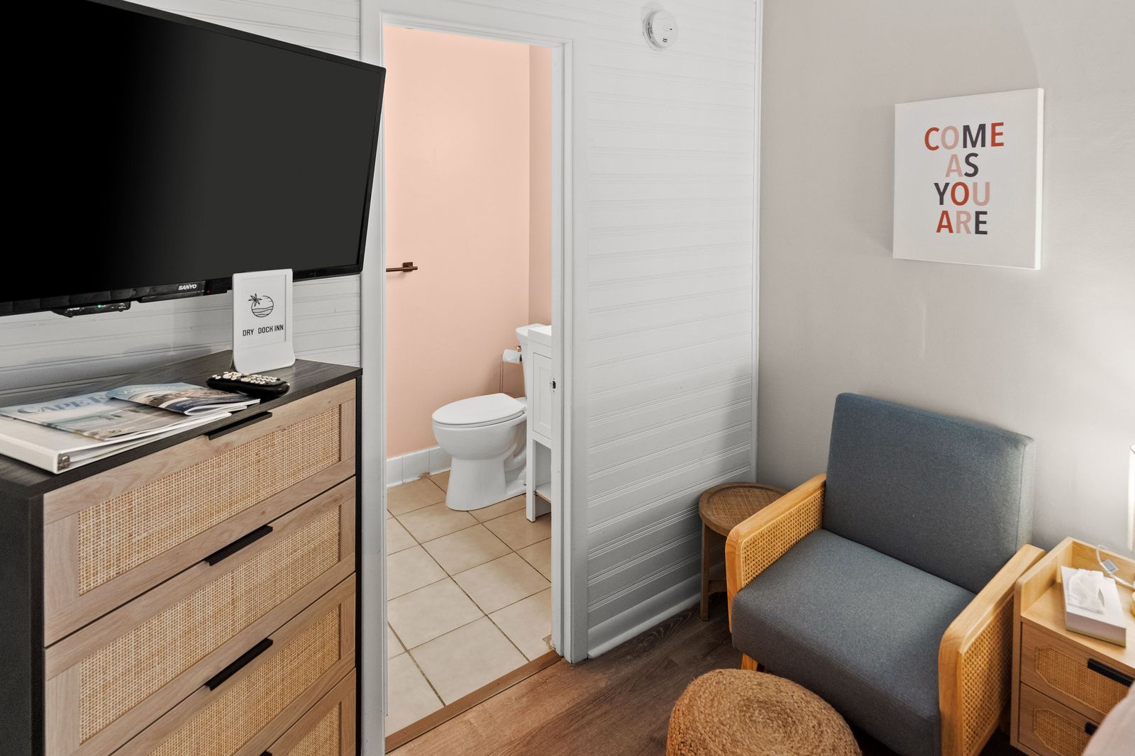 A hotel room with a TV, a chest of drawers, an armchair, and a bathroom visible through an open door.