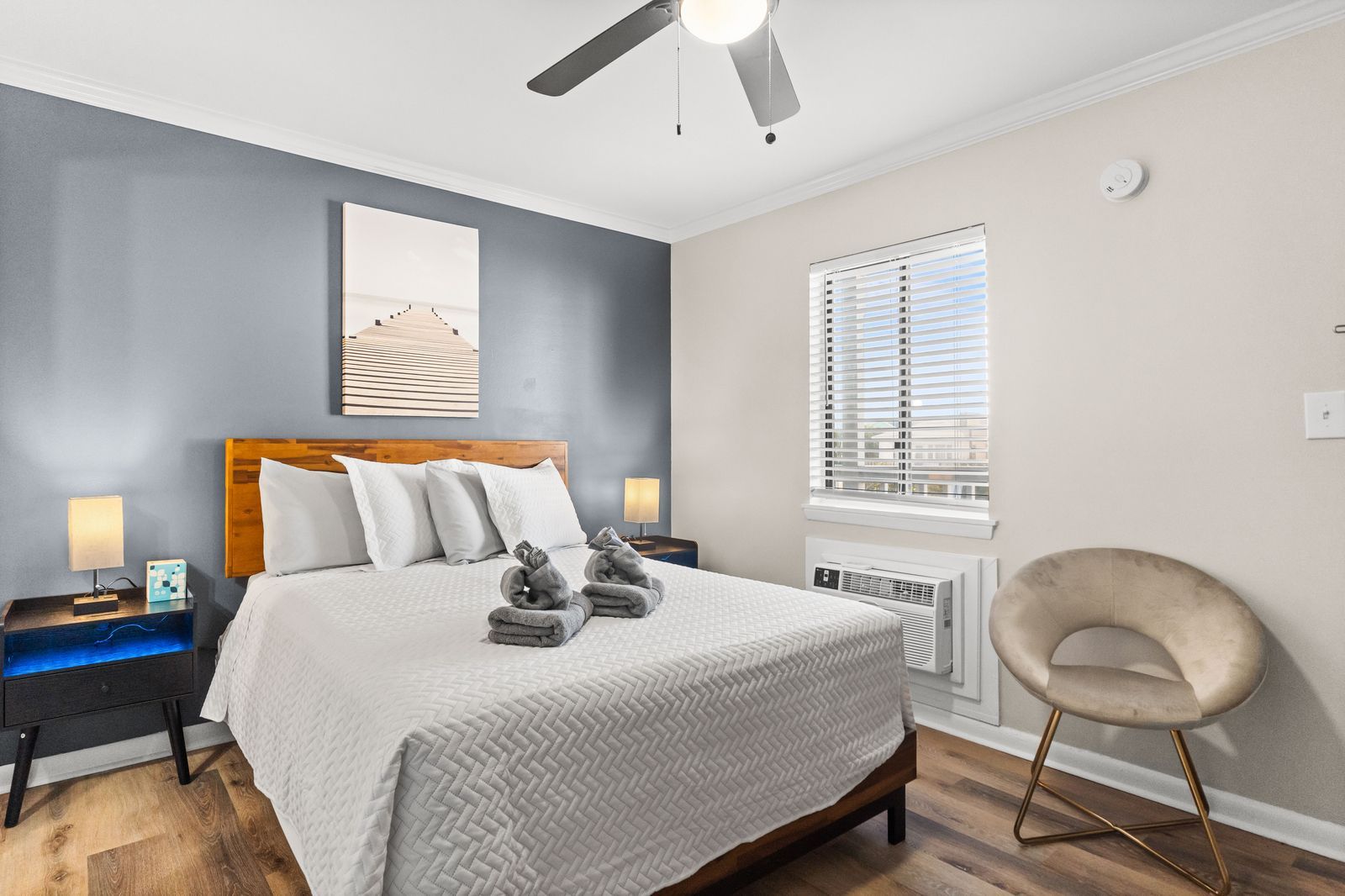 Bedroom with a queen bed, bedside tables, art, and a chair. One wall is gray, the others are light gray with a window.