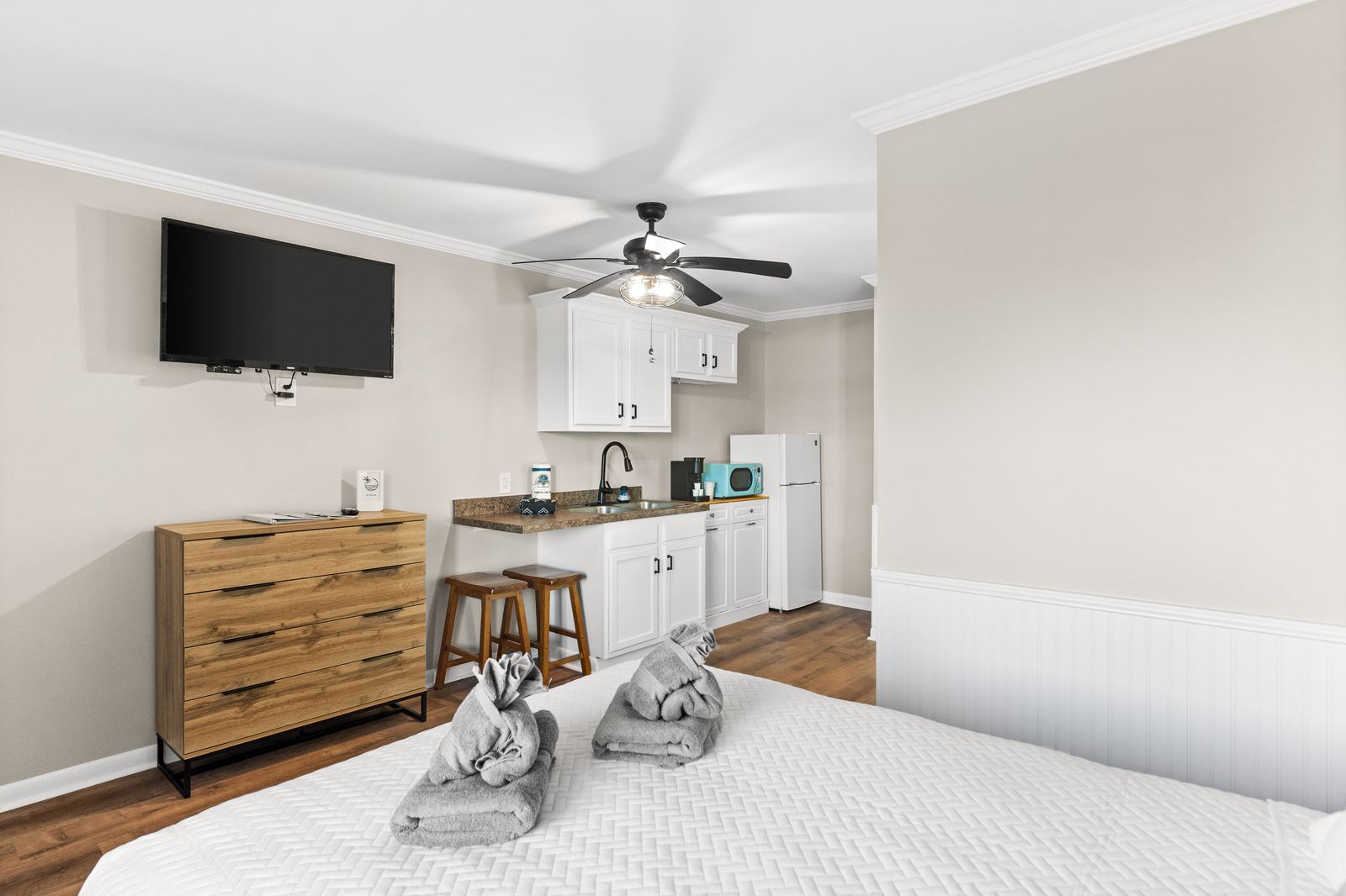Studio apartment with kitchenette, bed, TV, dresser, and ceiling fan.