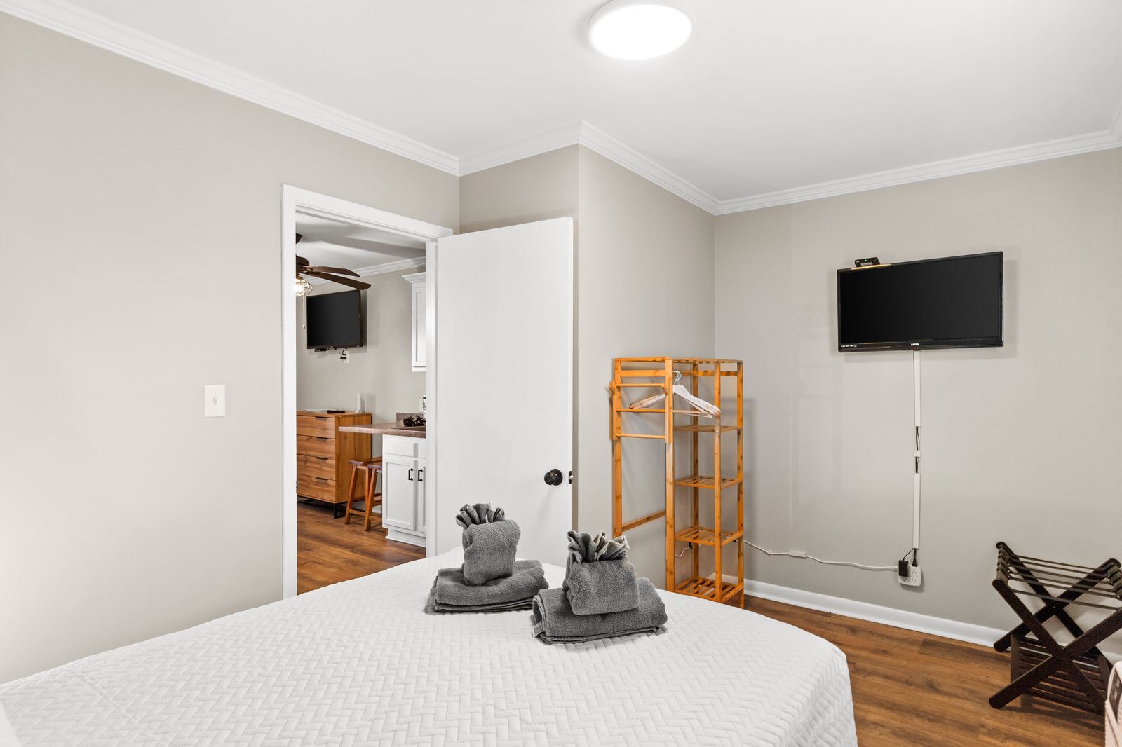 Bedroom with white bed, TV, wooden rack, and doorway leading to a kitchenette.