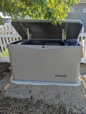 KOHLER outdoor generator with open lid in a gravel yard, shown beside a white fence.