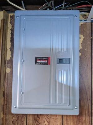 Gray electrical breaker panel mounted on a wooden wall, with wires visible above it.