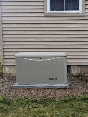 Outdoor beige Kohler generator beside a house with tan siding and a basement window