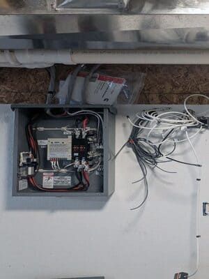 Open electrical junction box with exposed wiring and loose cables on a white wall under a countertop.