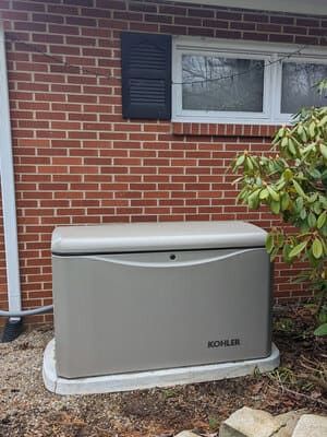 White KOHLER generator beside a brick house under a window with black shutters.