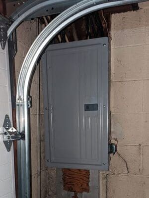 Gray electrical breaker box mounted on a cinderblock wall beside a metal garage door track.