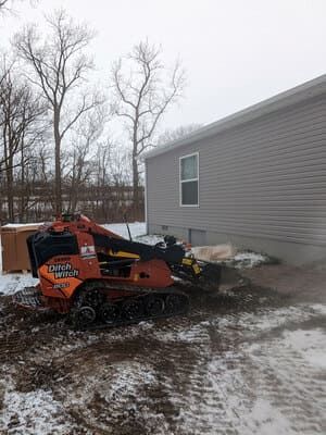Orange snow blower beside a gray house in a snowy yard with bare trees.