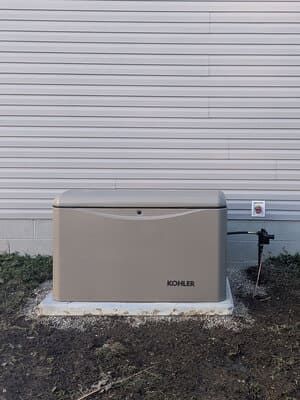 Beige Kohler generator beside a house wall and electrical meter on a gravel patch