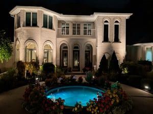 Large white mansion lit at night with a blue-lit pool and landscaped garden in front