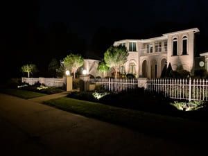 Lit mansion at night behind a white fence, with manicured landscaping and driveway in front.