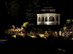 Illuminated white gazebo on a dark hillside at night, surrounded by trees and garden lights