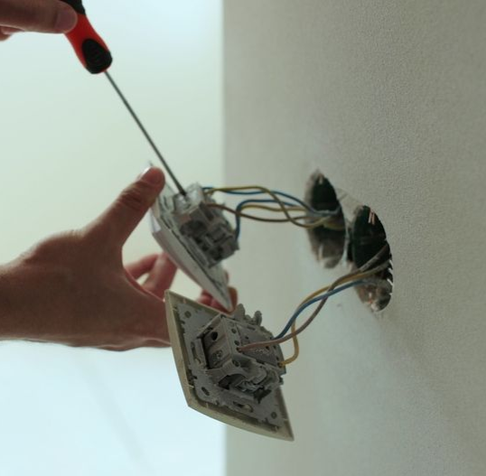 Hand turning a screwdriver to wire a wall switch with exposed electrical cables.